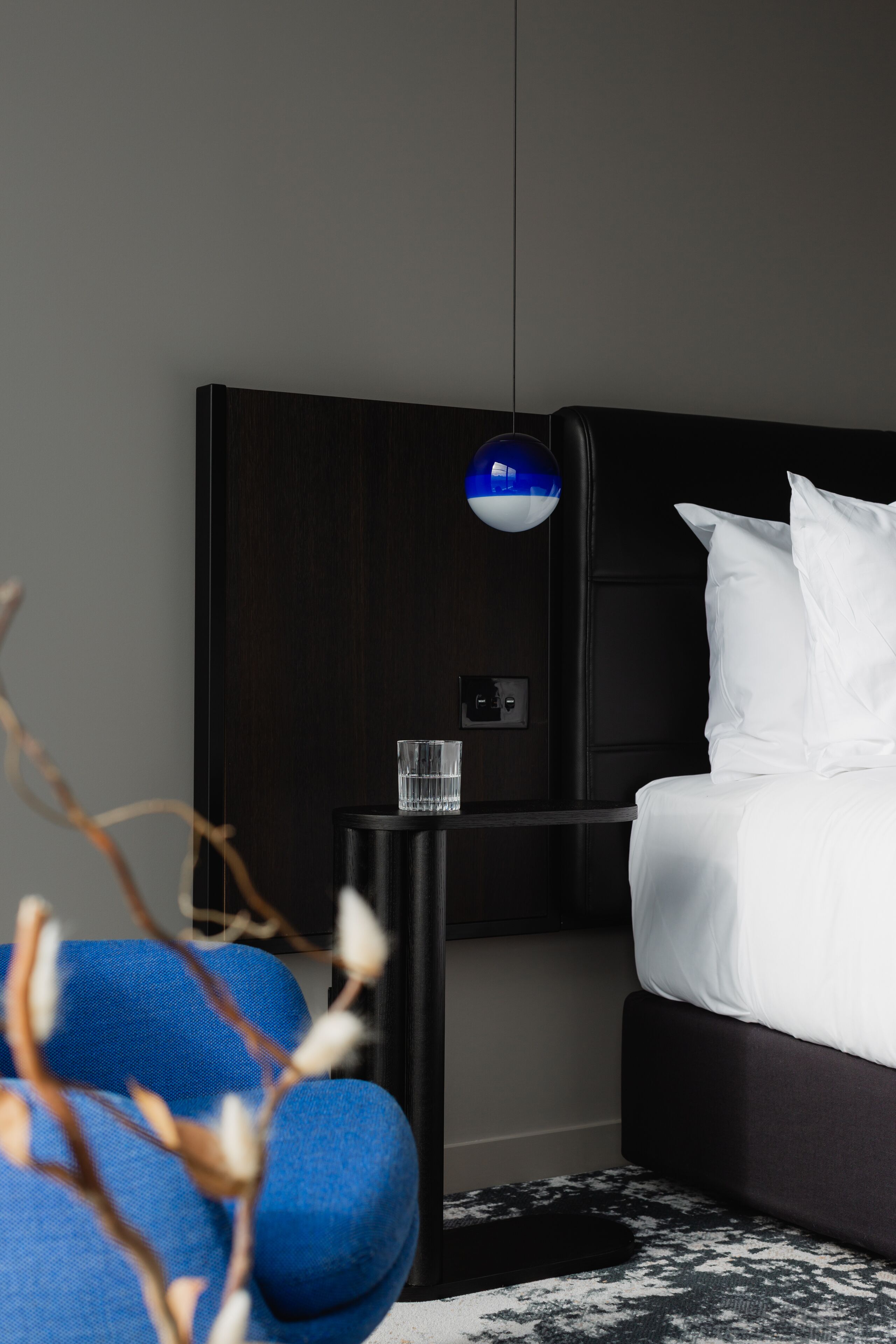 A modern hotel room with a dark wood headboard, a white bed, a small black nightstand with a glass, and a blue spherical light fixture.