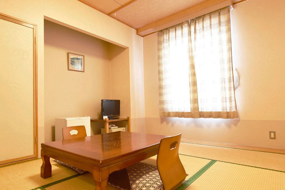 Ein Zimmer mit Tatami-Böden, einem Holztisch, einem Fernseher auf einem Schrank und einer Schiebetür.