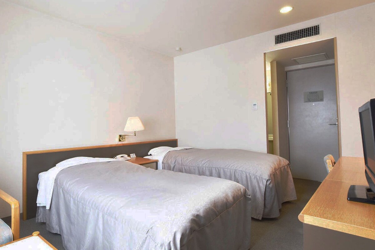 Ein Hotelzimmer mit zwei Betten, einem Schreibtisch, einem Stuhl, einer Lampe und einem Fernseher.