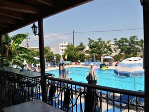 Zona de piscina con sillones de descanso y sombrillas, rodeada de vegetación y edificios.