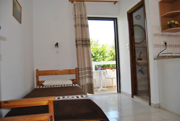 Un dormitorio con cama, una mesita, una silla, un baño con lavamanos y espejo, y un balcón con vistas a zonas verdes.
