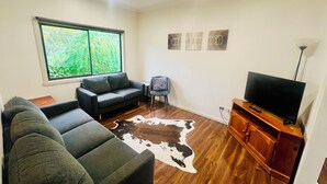 Ein Wohnzimmer mit einer Couch, einem Fernseher, einem Holzschrank, einem Kuhfellteppich und einem Fenster mit Blick ins Grüne.