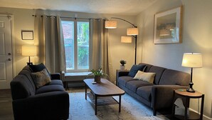 Sala de estar con dos sofás, una mesa de centro y una mesita auxiliar. Hay una ventana con cortinas, una puerta y un cuadro en la pared.
