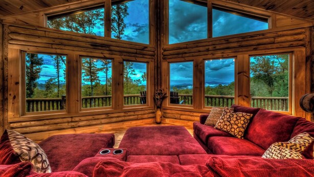 A log cabin interior with a large windowed corner, a red sofa, and a view of the forest. - Serenity Peak | Mountain View | Close To Town | Paved | Dog Friendly (Blue Ridge)