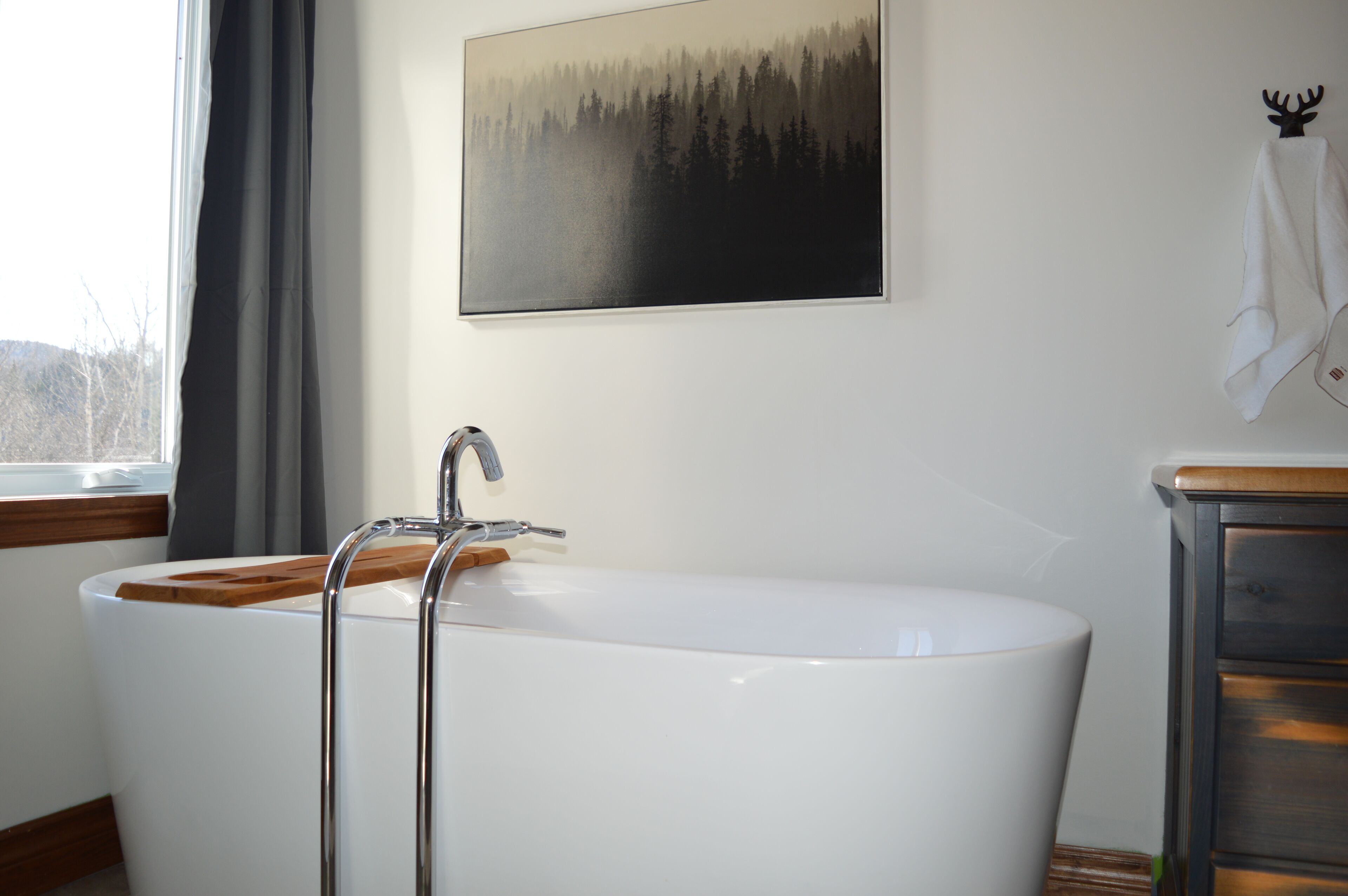 A modern bathroom with a freestanding bathtub, a wooden sideboard, and a forest-themed painting on the wall.