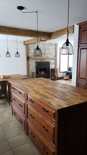 A spacious kitchen with a large wooden island, a stone fireplace, hanging pendant lights, and wooden cabinets.