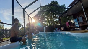 A pool area with a metal roof structure, surrounded by trees and a building with a balcony.