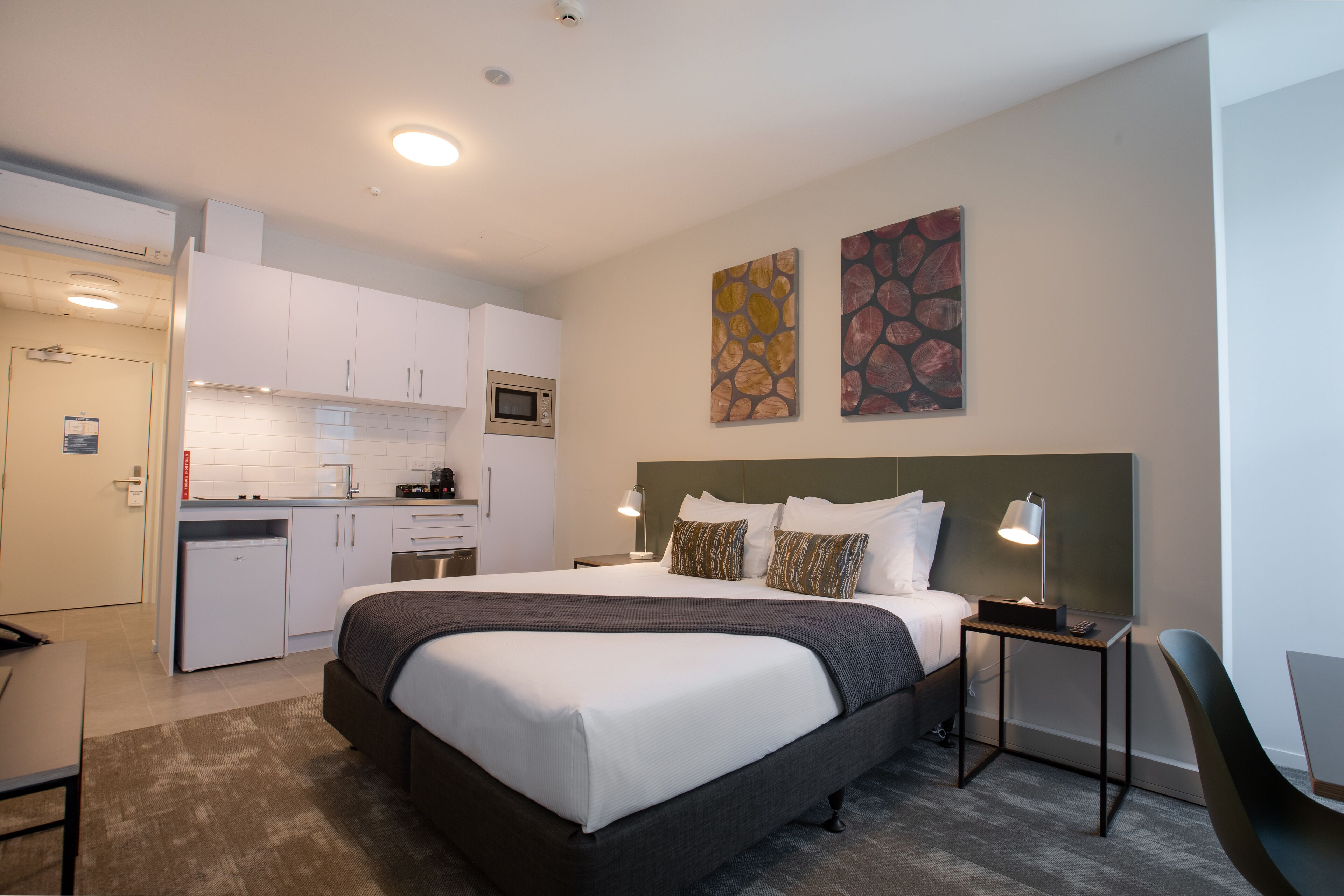 Habitación de hotel pequeña y funcional, con cama, mesitas de noche, escritorio, kitchenette y cuadros decorativos en las paredes.