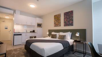 Habitación de hotel pequeña y funcional, con cama, mesitas de noche, escritorio, kitchenette y cuadros decorativos en las paredes.