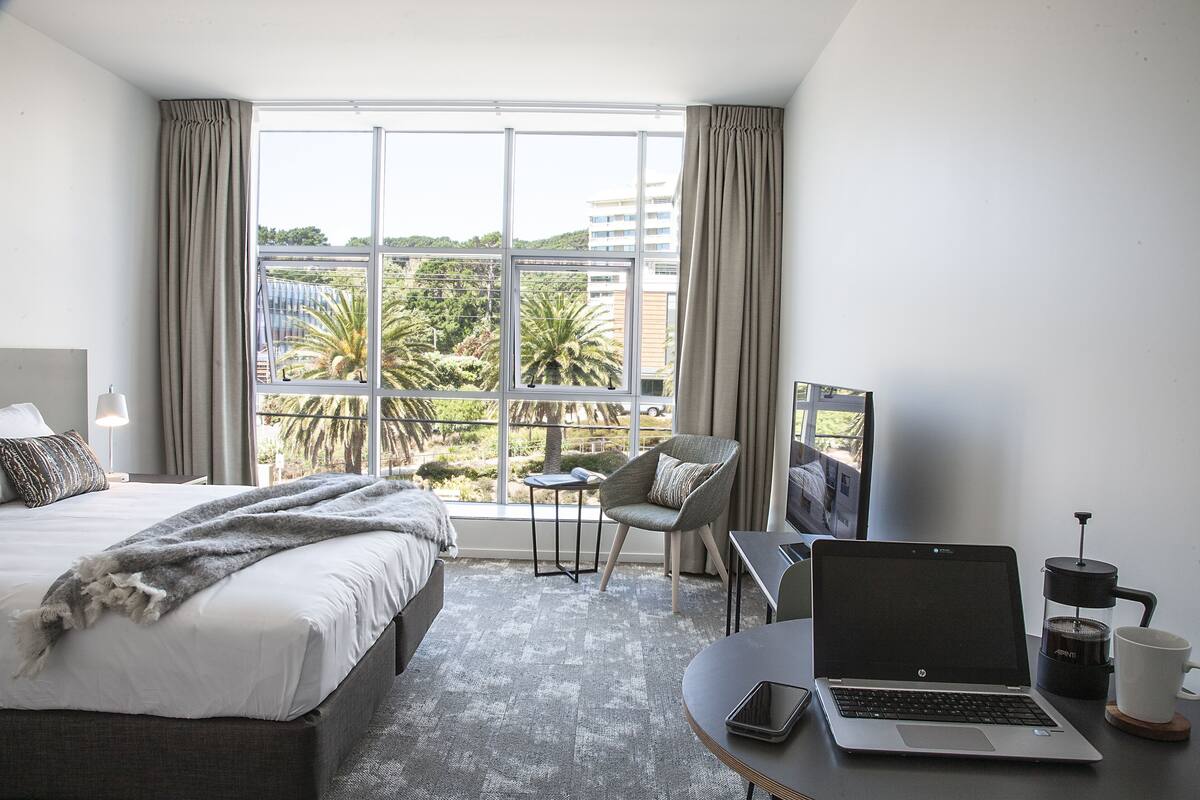 Habitación de hotel con cama, escritorio con computadora portátil, silla, cafetera y ventana grande con vista a palmeras y edificios.