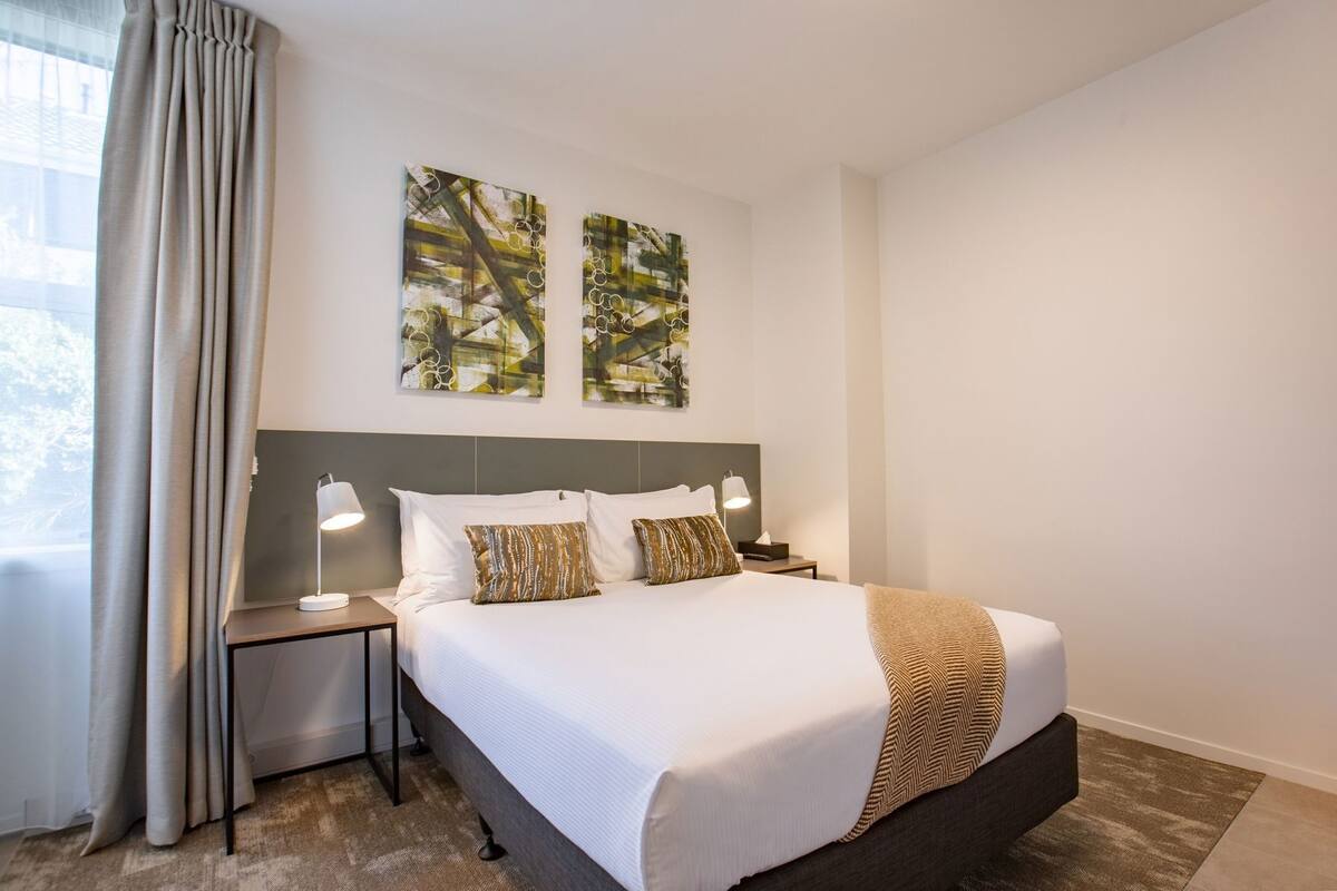 Habitación de hotel con cama, mesitas de noche, lámpara, dos cuadros abstractos y ventana con cortinas.