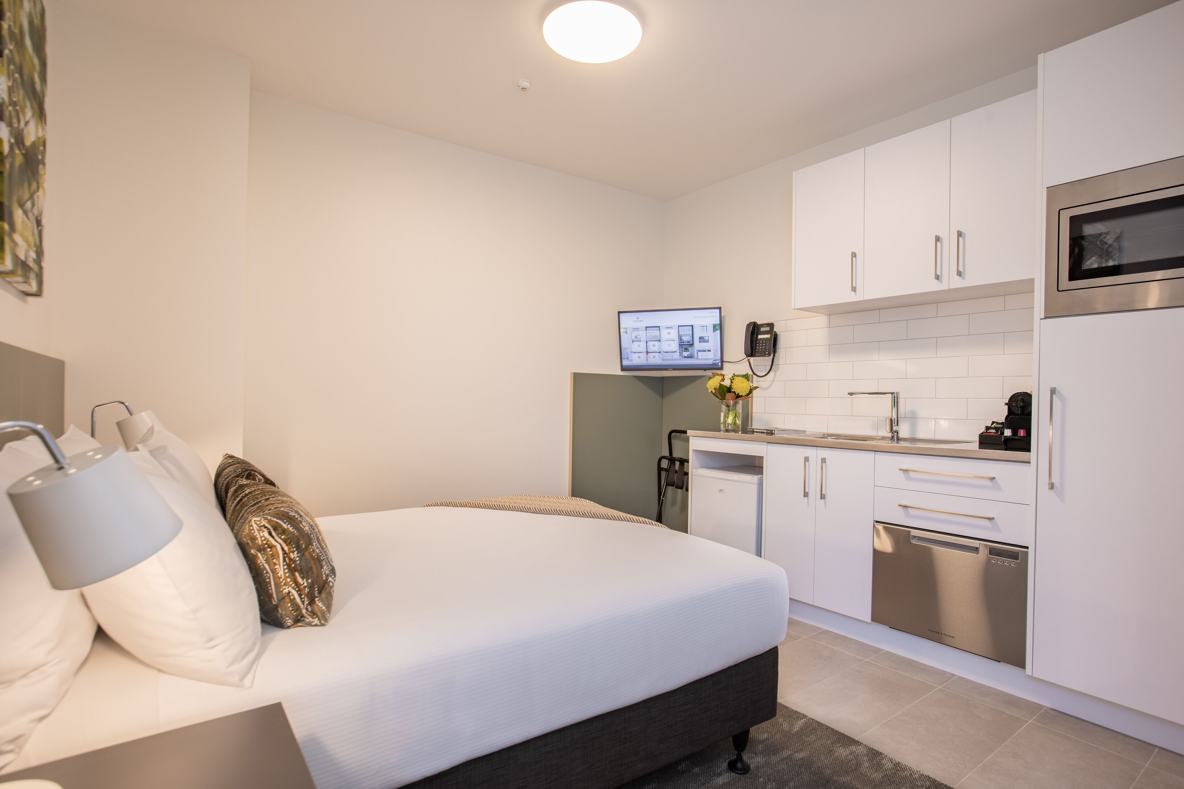 Una habitación de hotel moderna con cama, lámpara de noche y una pequeña kitchenette equipada con microondas y lavavajillas.