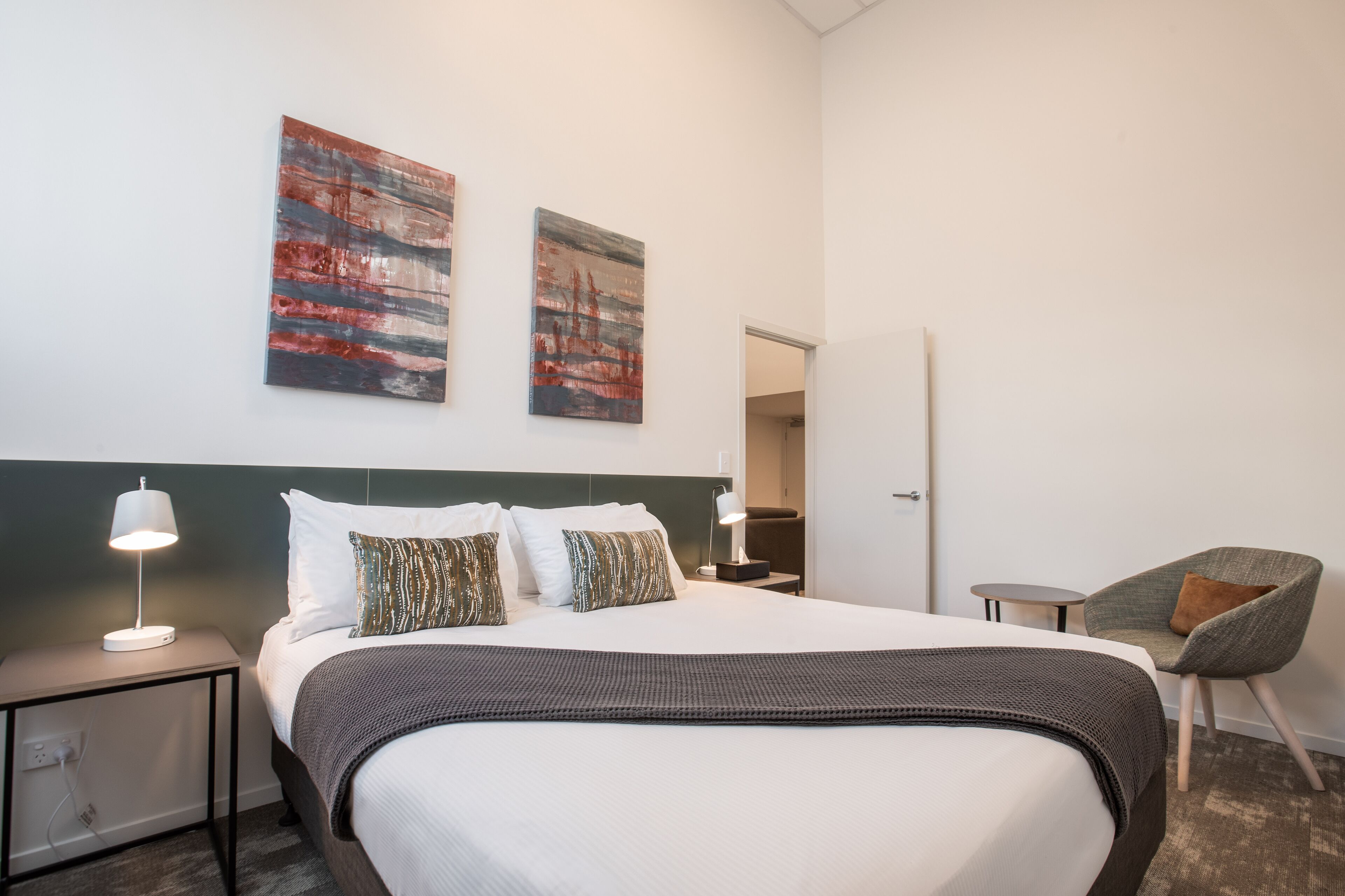A hotel room with a large bed, two bedside tables with lamps, a chair, and two abstract paintings on the wall.