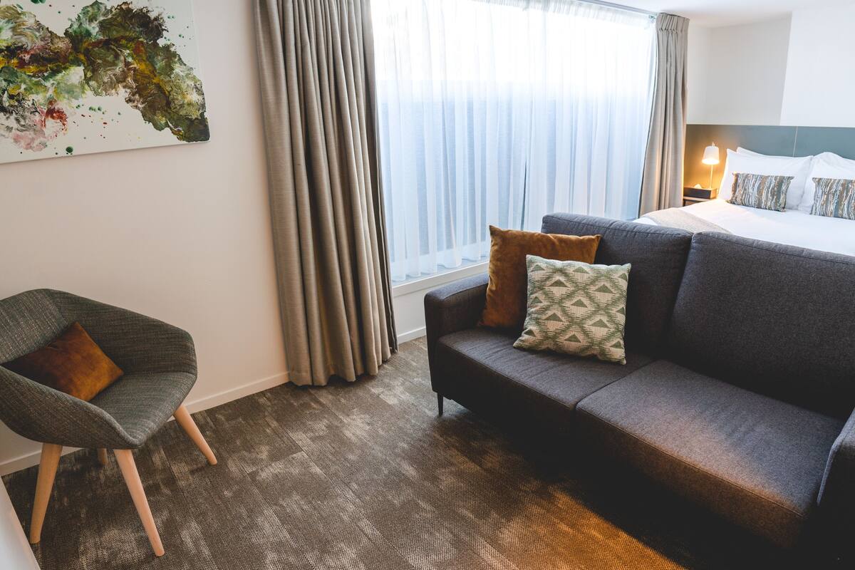 Habitación de hotel con un sofá gris, una silla, una cama con almohadones estampados, un cuadro en la pared y una ventana con cortinas.