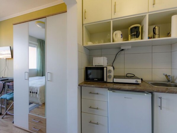 Een compacte keuken met een magnetron, broodrooster en een spoelbak. Daarbij een kleine koelkast en een kast met keukengerei. Een stoel en een bed zijn zichtbaar in de aangrenzende kamer.