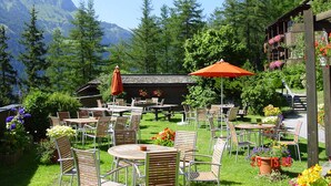 An outdoor seating area with tables and chairs, surrounded by greenery and flowers, with a mountain in the background. - Cosy apartment with own sauna in hotel (Matrei in Osttirol)