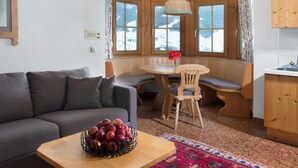 A cozy living room with a sofa, a wooden table with a fruit bowl, a patterned rug, and a bay window with a view of snow-covered mountains. - Cosy apartment with own sauna in hotel (Matrei in Osttirol)