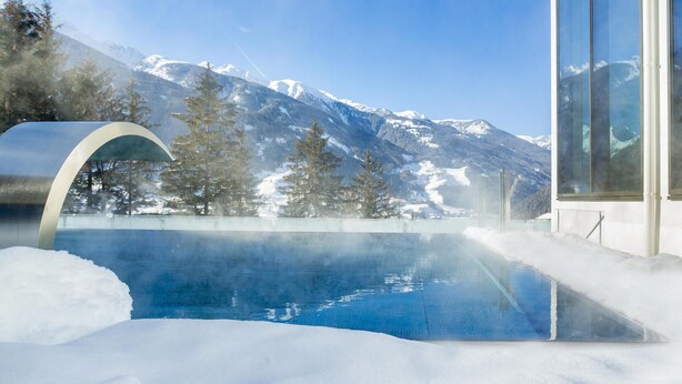 A modern building with large windows overlooking a pool with a snowy mountain backdrop. - Cosy apartment with own sauna in hotel (Matrei in Osttirol)