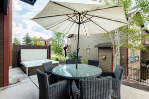 An outdoor patio with a glass table and wicker chairs, a large umbrella, and a hot tub.