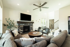 A living room with a stone fireplace, a ceiling fan, a flat-screen TV, and a staircase leading to another level.