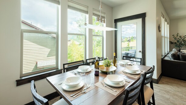 Un coin repas avec une table en bois dressée pour quatre personnes, de grandes fenêtres et une vue sur l’extérieur.