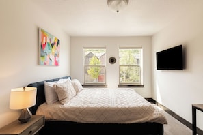 A bedroom with a bed, a nightstand, a lamp, a wall-mounted TV, and two windows with a view of houses and trees.