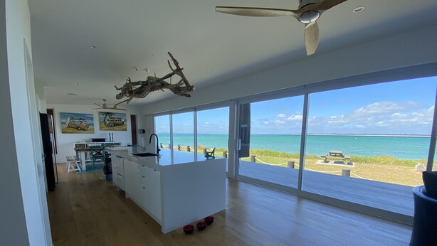 Una cocina amplia con una isla grande, un ventilador de techo y vistas a la playa a través de ventanas panorámicas.