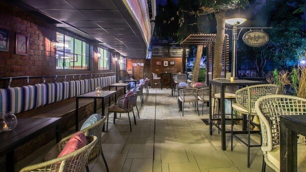 An outdoor restaurant with wooden tables and wicker chairs, a brick wall, and a sign reading "Red Bull." - T24 Retro (Mumbai)