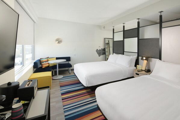 A hotel room with two beds, a small desk, a chair, a colorful striped rug, and a window with blinds. - Aloft by Marriott Lubbock (Lubbock)