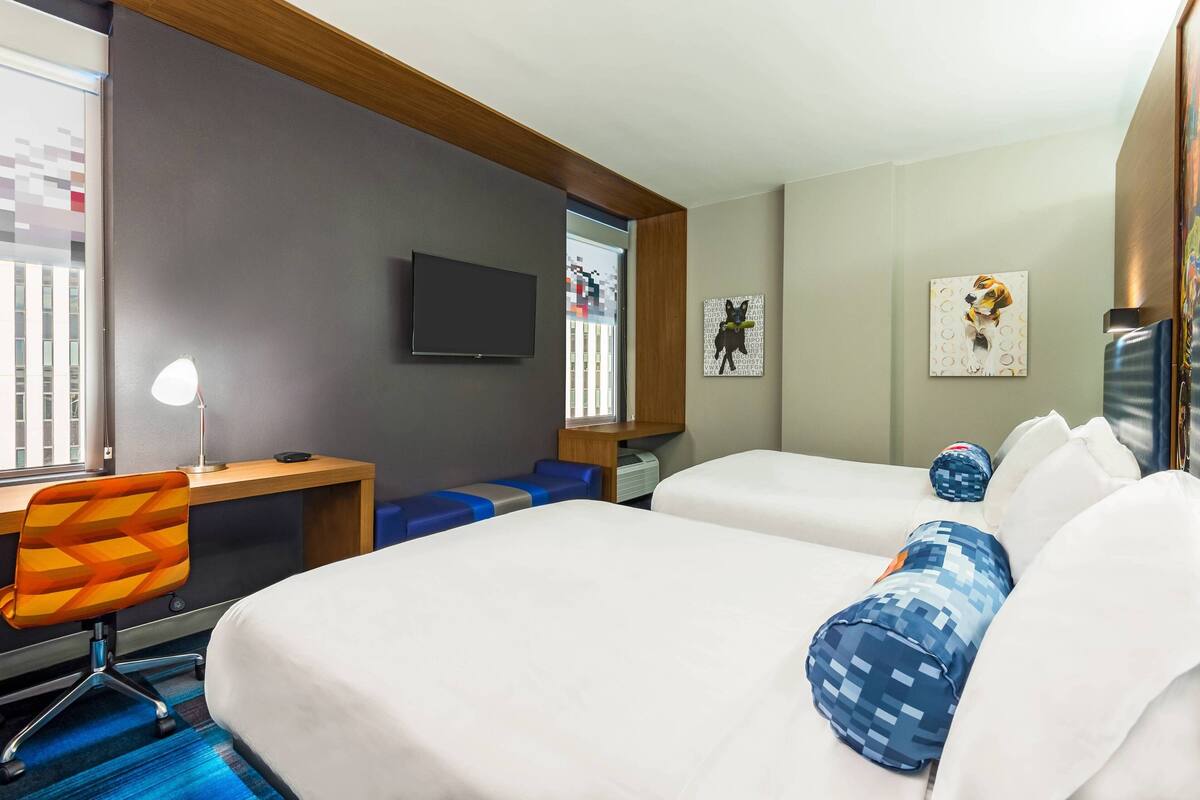 A hotel room with two beds, a desk with a chair, a TV, and artwork on the walls.