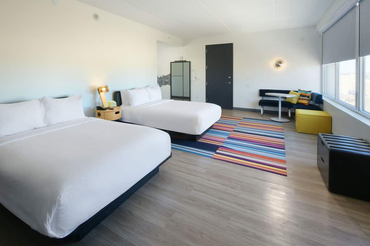 Una habitación de hotel moderna con dos camas, una pequeña mesa, un sofá y un ventanal grande.