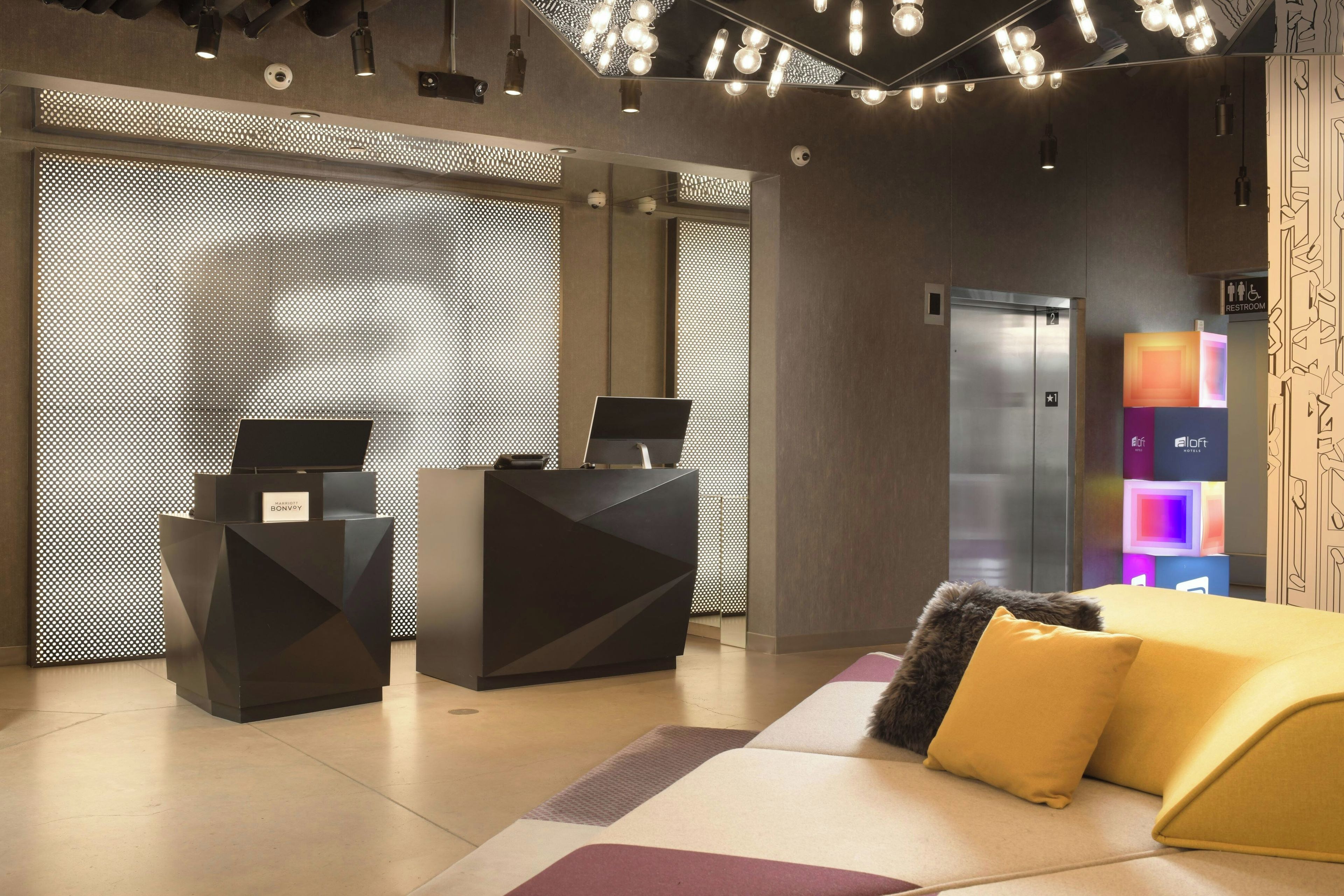 A hotel reception area with a modern design, including a reception desk, a digital screen, and a seating area with a sofa and cushions.