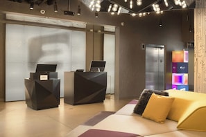 A hotel reception area with a modern design, including a reception desk, a digital screen, and a seating area with a sofa and cushions.