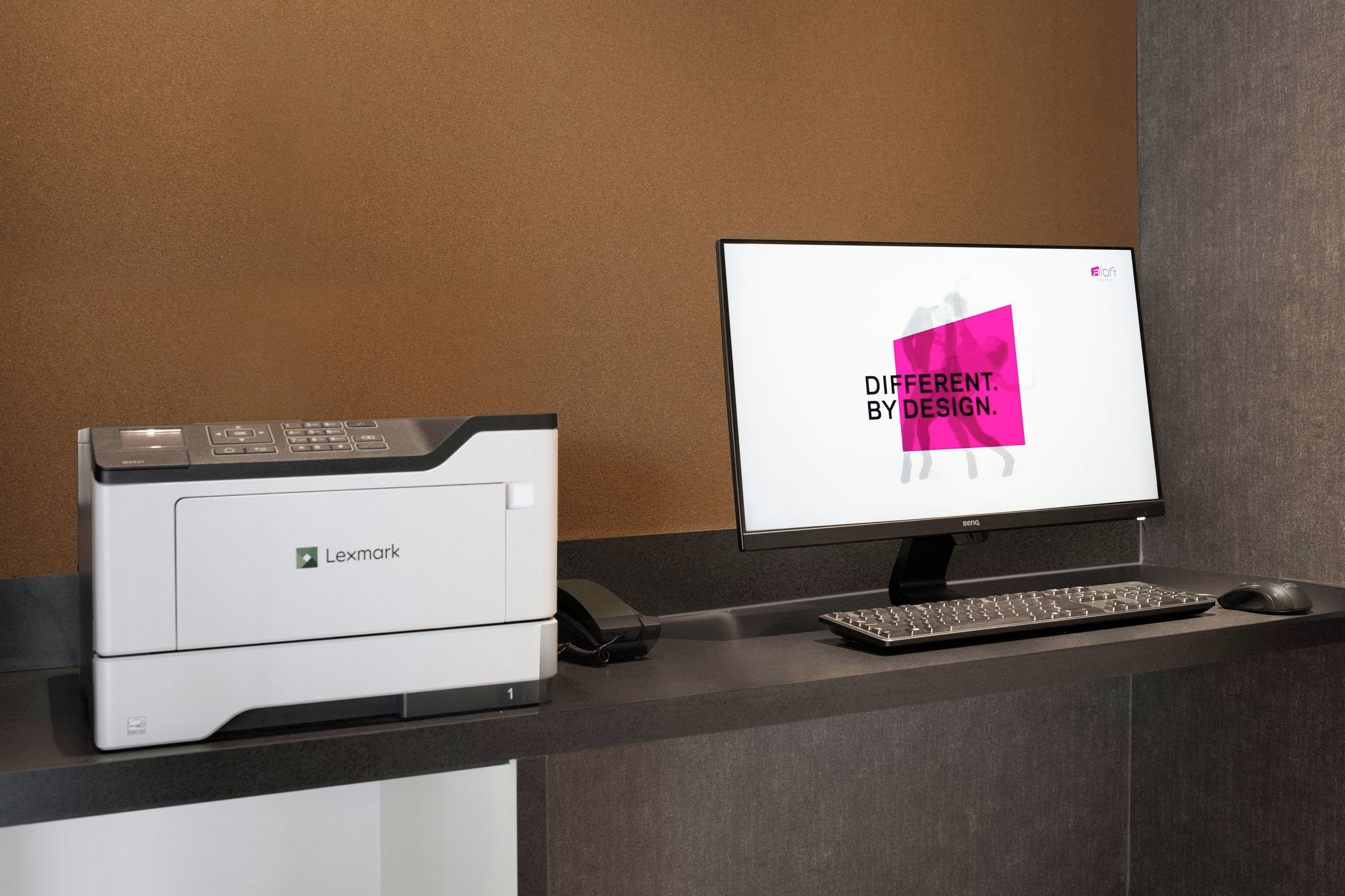 A printer, a monitor, and a keyboard on a desk with a corkboard wall.