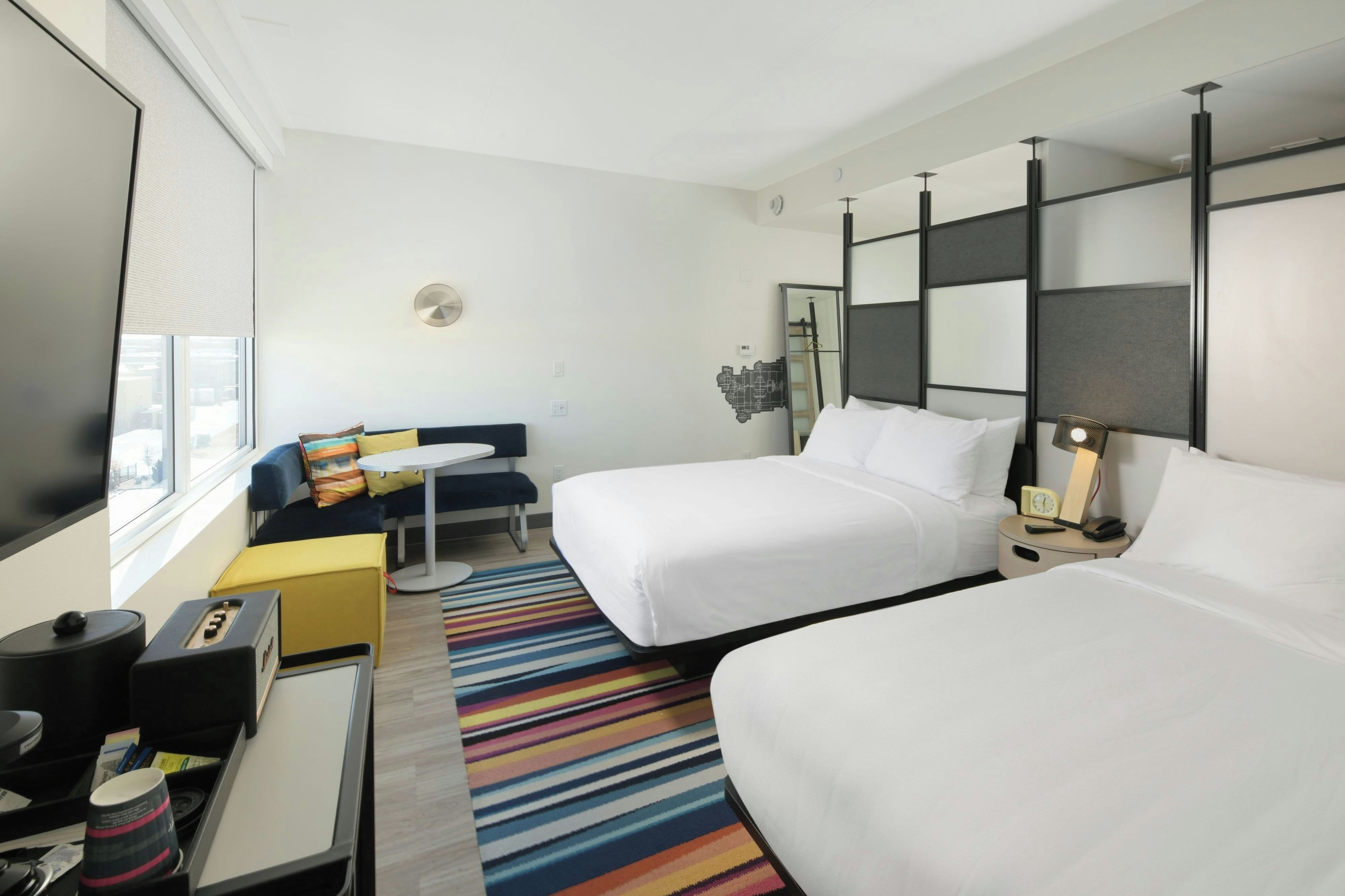 A hotel room with two beds, a desk, a chair, a small table, a colorful striped rug, and a window with blinds.