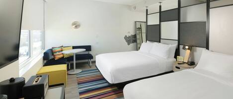 A hotel room with two beds, a desk, a chair, a small table, a colorful striped rug, and a window with blinds.