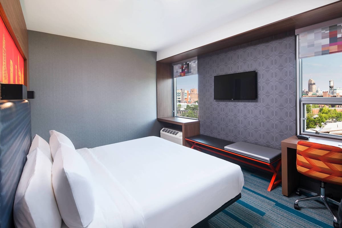 Habitación de hotel con cama, escritorio, silla, ventana con vistas a la ciudad y televisor de pared.