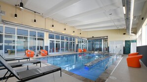 An indoor swimming pool with clear glass walls, orange chairs, and a modern ceiling design. - Aloft by Marriott Lubbock (Lubbock)