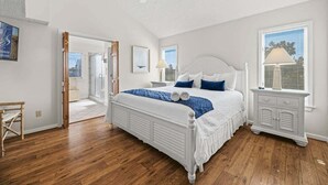 Un dormitorio con cama blanca, ventilador de techo, mesita de noche, ventana con persianas y suelo de madera.
