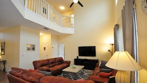 A spacious living room with a high ceiling, a staircase, a flat-screen TV, leather sofas, and a lamp. - The Lake House! 7Br 4.5ba with private pool! (Kissimmee)