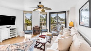 A spacious living room with a large flat-screen TV, comfortable seating, a ceiling fan, and large windows offering a view of the outdoors.