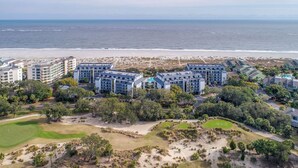 An aerial view of a coastal hotel complex with multiple buildings, a golf course, and a beach in the background.