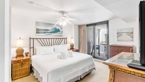 A hotel room with a bed, a ceiling fan, a painting of a wave, a desk with a keyboard, and a view of the outdoors through a glass door.