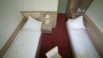 A small, compact hotel room with two single beds, a small bedside table with a lamp, and a red carpeted floor.