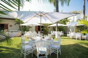 An outdoor dining area with round tables and white chairs, shaded by large umbrellas, surrounded by palm trees and a building. - Gamma Cuernavaca Puerta Paraiso (Cuernavaca)