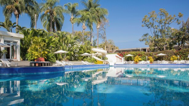 A swimming pool with lounge chairs and umbrellas, surrounded by palm trees and a building. - Gamma Cuernavaca Puerta Paraiso (Cuernavaca)