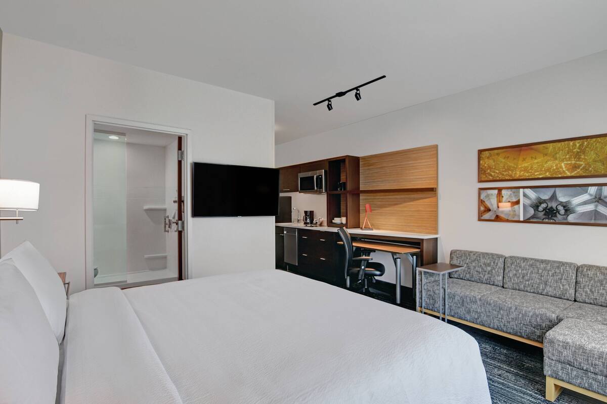 Una habitación de hotel moderna con una cama grande, un escritorio con computadora, un sofá, un televisor de pantalla plana montado en la pared y un microondas en la kitchenette.