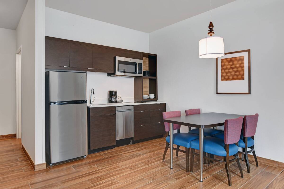 Una cocina moderna con un refrigerador, un microondas y un lavavajillas de acero inoxidable. Hay una mesa de comedor con sillas azules y una lámpara colgante arriba.