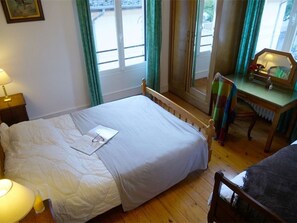 Un dormitorio con cama, escritorio y silla. Se ve una ventana con cortinas, una lámpara en la mesita de noche y un cuadro enmarcado en la pared.