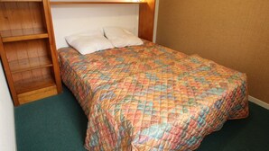 Una habitación pequeña con una cama individual, cabecera de madera, colcha colorida y estantería.
